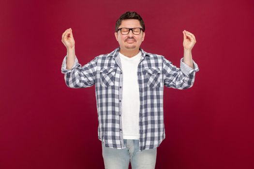 Portrait of handsome middle aged man in casual checkered shirt and eyeglasses standing with raised arms in yoga pose, doing meditating and relaxing. indoor studio shot, isolated on dark red background