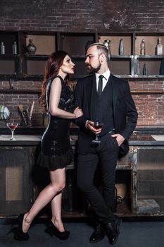Flirt on a party. Girl holding glasses of vine and looking with passion on bearded man. Studio shot, brick wall