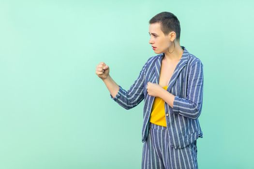 Profile side view portrait of serious beautiful short hair young stylish woman in casual striped suit standing with boxing fists and looking at camera. studio shot isolated on light green background.
