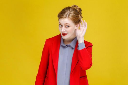Ginger red head woman in red suit listening secret. Studio shot, isolated on yellow background