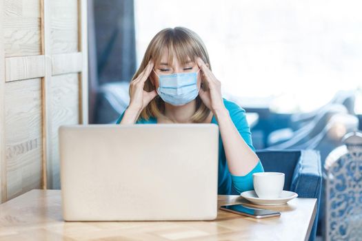 Headache or confusion. Portrait of young woman with surgical medical mask is sitting and working on laptop and holding her painful head or thinking. Indoor working, medicine and health care concept.