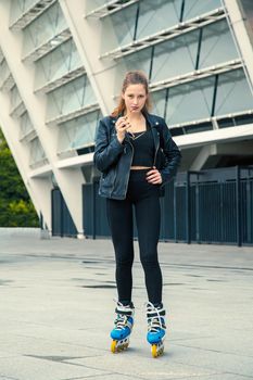 Girl on rollerblades standing in building background. Young fit women girl in blue sunglasses, jeans and jacket on roller skates riding outdoors after rain.