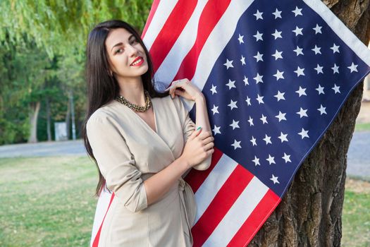 Beautiful young woman with classic dress near american flag in the park. fashion model holding us smiling and looking at camera. usa lifestyle. posing near tree and toothy smile.