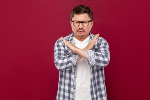There is no way. Portrait of serious handsome middle aged business man in casual checkered shirt and eyeglasses standing with X sign and looking at camera. studio shot, isolated on dark red background