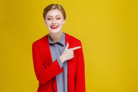 Redhead business woman in red suit showing at copy space. Studio shot, isolated on yellow background