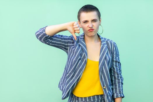 Dislike. Portrait of dissatisfied beautiful short hair young stylish woman in casual striped suit standing with thumbs down and looking at camera. indoor studio shot isolated on light green background