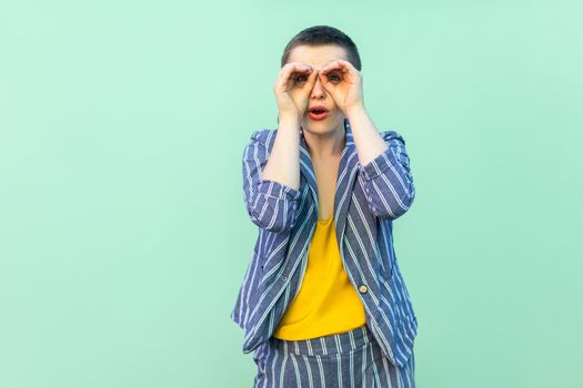 Portrait of amazed handsome beautiful short hair young stylish woman in casual striped suit standing with binoculars hands on eyes surprised. indoor studio shot isolated on light green background.