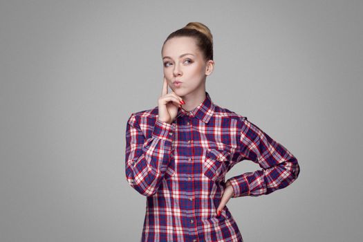 Thoughtful beautiful blonde girl in pink, red checkered shirt, collected bun hairstyle standing and looking away and and thinking touching face . indoor studio shot. isolated on gray background