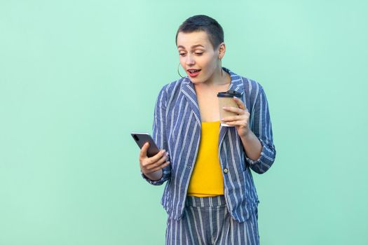 Portrait of excited beautiful with short hair young blogger woman in striped suit standing, reading interesting news and drink coffee, looking at phone. Indoor, isolated, studio shot, green background