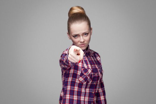 Hey you! beautiful blonde girl in pink checkered shirt, collected bun hairstyle, makeup standing and looking and pointing at camera with serious face. indoor studio shot. isolated on gray background