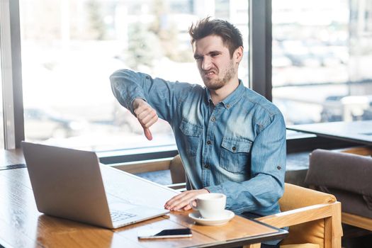 Dislike! Side view portrait of negative critique bearded young freelancer in blue jeans shirt are sitting in cafe and making video call on laptop and showing thumb down to his companion. indoor,inside