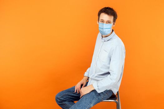 Portrait of young worker man with surgical medical mask sitting and looking at camera with serious face. health care and medicine concept. indoor studio shot isolated on orange background.