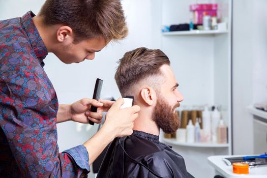 Handsome man with fashion highlighted hair wearing white shirt doing a haircut for man with brown hair at barber shop.