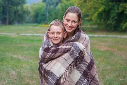 Young sister and brother with freckles on their faces stand covered with plaid in park, get warm, smiling and looking at camera.