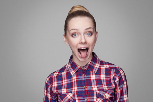 portrait of funny beautiful blonde girl in pink checkered shirt, collected bun hairstyle and makeup standing with open mouth, tongue out and big eyes. indoor studio shot. isolated on gray background