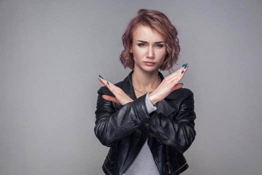 Portrait of serious woman with short hairs and makeup in casual style black leather jacket standing making X sign with her arms to stop doing something. indoor studio shot, isolated on grey background