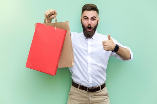 Online shopping. Young adult businessman thumbs up and looking at camera with surprised face. Indoor, studio shot on light green background