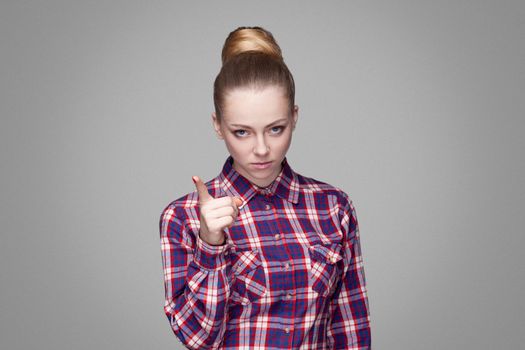 serious beautiful blonde girl in red, pink checkered shirt, collected bun hairstyle, makeup standing and looking at camera with warning sign. indoor studio shot. isolated on gray background