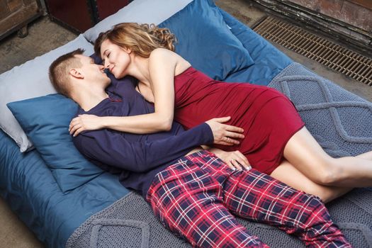 Portrait of happy young family sleeping in the bedroom, in sleepwear lying on bed with blue knitting plaid enjoying each other. Husband with love touching his future baby. Indoor, studio shot