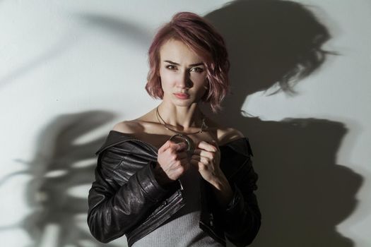 beautiful woman with makeup and short pink hair in grey dress and black leather jacket posing on grey background with dark shadow of leaf texture pattern. indoor studio shot.