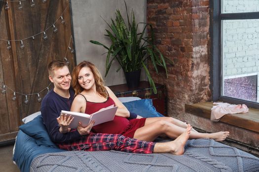 Portrait of happy young family have a rest in the bedroom, in sleepwear holding album are talking and smiling while spending time together in bed with blue plaid. Indoor,studio shot, looking at camera