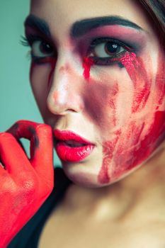 portrait of freak monster with mess dirty colored makeup on her face. crying woman with red bloody tears and hand. halloween concept on green background. studio shot, dark brown eyes.