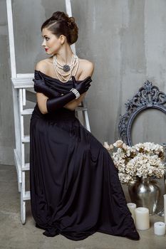 Beautiful young fashion caucasian model in black long dress and fashion makeup and beautiful hairdo posing sitting on the chair and looking at camera. Studio shot.