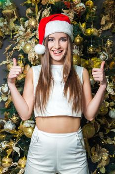 Young funny beautiful fashion model with dark eyes, brown hair and santa hat celebrating new year at home. new year decoration, with positive emotion posing and looking at camera. yellow gold decor..