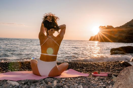 Young woman in swimsuit with long hair practicing stretching outdoors on yoga mat by the sea on a sunny day. Women's yoga fitness pilates routine. Healthy lifestyle, harmony and meditation concept.