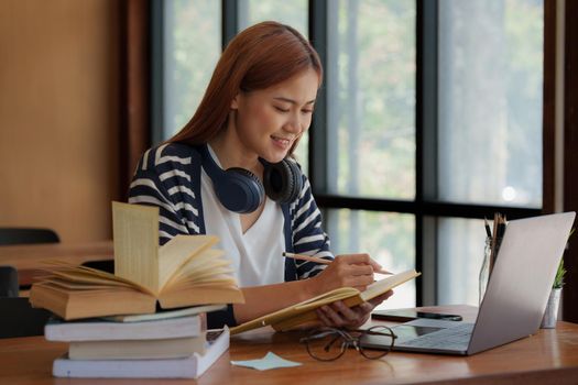 Young asian girl student wear headphone and writing a notebook and watching online class by laptop. e-learning education concept