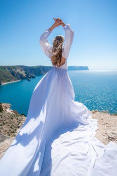 Blonde with long hair on a sunny seashore in a white flowing dress, rear view, silk fabric waving in the wind. Against the backdrop of the blue sky and mountains on the seashore