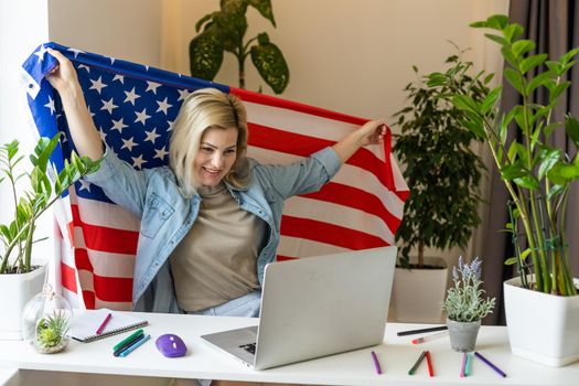 Happy woman employee sitting wrapped in USA flag, shouting for joy in office workplace, celebrating labor day or US Independence day. Indoor studio studio shot isolated on yellow background.