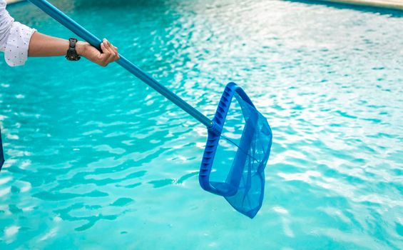 A man cleaning pool with leaf skimmer. Man cleaning the pool with the Skimmer, Person with skimmer cleaning pool, Hands holding a skimmer with blue pool in the background