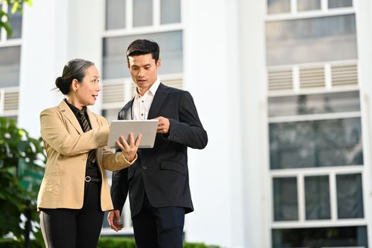 Senior female entrepreneur and young businessman discussing work on tablet while standing outdoor with modern office building in background.