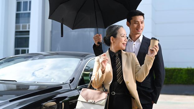 Smiling mature businesswoman and her assistant standing by black car at parking with modern office building in background.