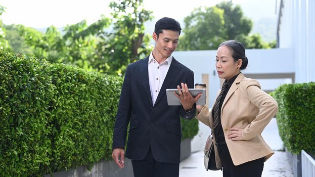 Young businessman and mature businesswoman discussing work with tablet device while walking outside office building.