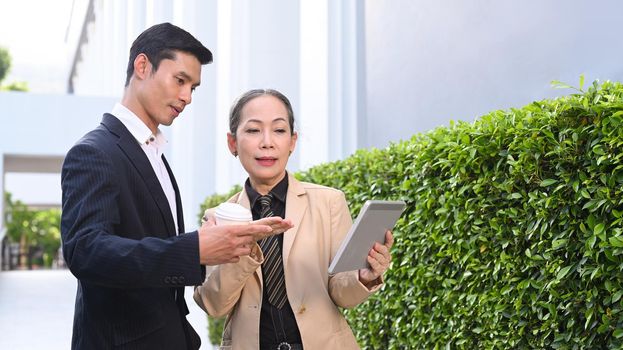 Mature businesswoman and young businessman discussing work with digital tablet while walking outdoors at the city streets.