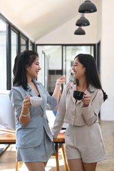 Two cheerful young woman office workers talking each other during coffee break.