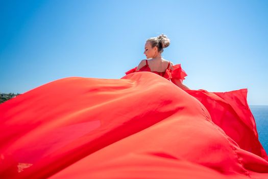 Blonde with long hair on a sunny seashore in a red flowing dress, back view, silk fabric waving in the wind. Against the backdrop of the blue sky and mountains on the seashore