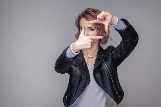 Portrait of beautiful girl with short hairstyle and makeup in casual style black leather jacket standing with cropping composition gesture and looking. indoor studio shot, isolated on grey background.