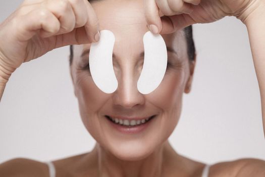 Patch Under Eyes. Beautiful middle aged woman with perfect skin showing white hydrogel patches with lifting anti-wrinkle collagen effect, gray background, indoor, studio shot, copy space, isolated