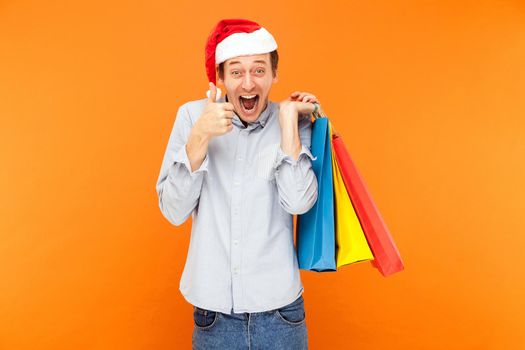 Thumbs up, like sign. Man shopaholic in red cap, holding many colorful bags after christmas shopping. Thumbs up and toothy smile. Indoor, studio shot on orange background