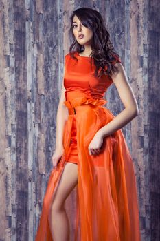 Young brunette woman in stylish orange silk dress posing on wooden background
