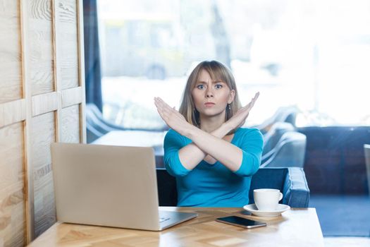 Enough! Portrait of warning aggressive young girl with blonde hair in t-shirt blouse are sitting in cafe and working on laptop and showing crossing raised arms like stop gesture. Indoor
