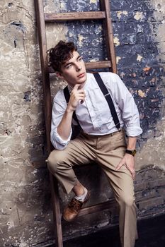 fashionable man in white shirt and beige pants with suspenders sitting and posing on wooden ladder on brick wall background. indoor, studio shot.