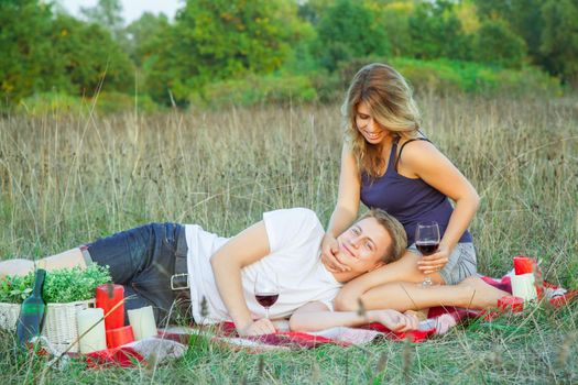 Beautiful young happy loving couple on picnic lying down on plaid in field on sunny summer day enjoying, holding and drinking wine and resting. looking at camera and smiling.