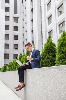 Attractive young businessman with laptop and coffee in hands on office building background. holding coffee and looking at display.