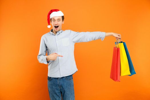 Amazement young adult man looking at camera with surprised face. Shocked man holding many colorful bags and pointing fingers on them. Studio shot. Orange background