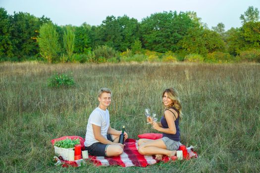Beautiful young happy loving couple on picnic lying down on plaid in field on sunny summer day enjoying, holding wine and resting. looking at camera and smiling.