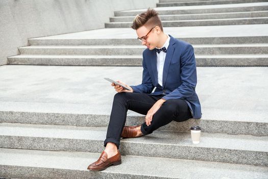 Attractive young businessman sitting with tablet and coffee in hands on office building background. holding tablet and looking at display with toothy smile.